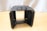 A black, rectangular heat sink with multiple fins is placed on a light wooden floor. The top part has a handle-like structure. The background is a textured grey fabric or curtain.