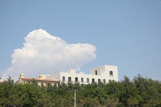 A white, rectangular building with an arched window design sits behind a row of green trees. Above the building, a large and fluffy white cloud is set against a clear blue sky. The architecture features clean lines and a modern aesthetic.