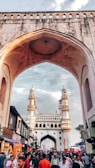 A historical archway frames a bustling street scene with a mix of people walking and conversing. In the background, a grand monument with four minarets rises under a partly cloudy sky. The architecture is intricate, and the street is lined with various shops and stalls.