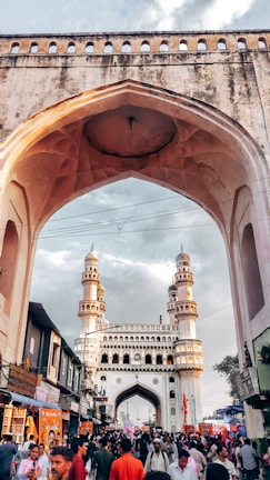 A historical archway frames a bustling street scene with a mix of people walking and conversing. In the background, a grand monument with four minarets rises under a partly cloudy sky. The architecture is intricate, and the street is lined with various shops and stalls.