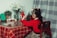 A child wearing a red sweater is decorating a small Christmas tree placed on a plaid-covered table. The table holds a red lantern, a miniature village set, and some flowers in a vase. The setting gives a cozy, festive vibe with holiday decorations.