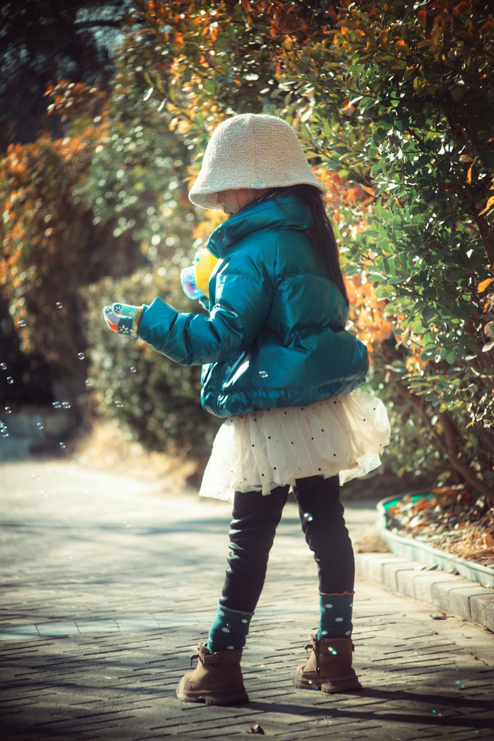 a little girl in a blue jacket and hat blowing bubbles