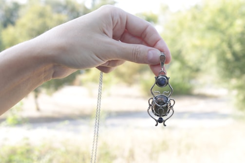 A hand is holding a pendant with an intricate design, featuring metalwork and stone details. The pendant is being displayed against a soft-focus, nature-filled background with green and brown tones.