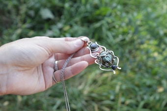 A hand is holding an intricately designed piece of jewelry, featuring a pendant with wire floral motifs and a green stone. The background is a blurred patch of greenery, suggesting an outdoor setting.