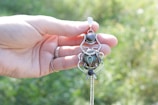 A hand holding an intricately designed silver pendant featuring a central round stone and ornamental elements. The background is softly blurred with greenery, creating a natural outdoor setting. The person is wearing a ring on one of their fingers.