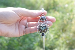 A hand holding an intricately designed silver pendant featuring a central round stone and ornamental elements. The background is softly blurred with greenery, creating a natural outdoor setting. The person is wearing a ring on one of their fingers.