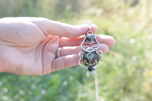 A hand with silver rings holds a pendant with ornate silverwork and dark gemstones, set against a blurred background of greenery.