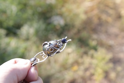 A hand holding an intricately designed piece of metallic jewelry, possibly a pendant, with a rounded central stone. The background is blurred greenery, suggesting an outdoor setting.