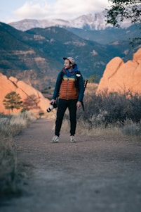 A person stands on a path in an outdoor setting, surrounded by rugged, rocky terrain with distant mountains and a cloudy sky. They are dressed warmly in a jacket and hold a camera with a long lens, suggesting they are hiking or exploring a natural area.