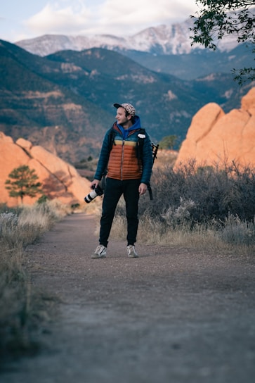 A person stands on a path in an outdoor setting, surrounded by rugged, rocky terrain with distant mountains and a cloudy sky. They are dressed warmly in a jacket and hold a camera with a long lens, suggesting they are hiking or exploring a natural area.