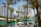 A tropical outdoor setting featuring tall palm trees and modern buildings in the background. There are green umbrellas and white lounging chairs, suggesting a resort or hotel area. The atmosphere is sunny and vibrant.