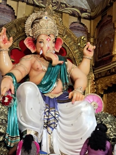 A large, intricately detailed statue of Lord Ganesha, adorned with a golden crown and multiple pieces of jewelry. The deity has a turquoise garment draped over one shoulder and is surrounded by ornate decorations. Two people stand in front, offering their respect.