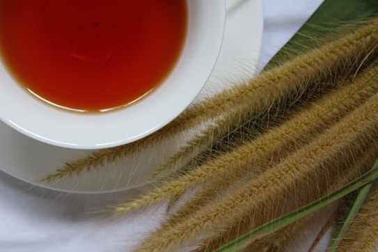 A white cup filled with reddish-brown tea is placed on a white surface. Next to the cup are several stalks of dried wheat or grass. The arrangement is simple, emphasizing a natural and rustic aesthetic.
