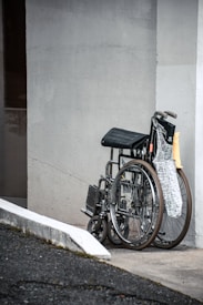 A wheelchair is parked against a light-colored wall next to a curb ramp. It is accompanied by a folded newspaper on the seat, and the scene conveys an outdoor setting with a partial view of the street.