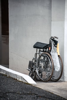 A wheelchair is parked against a light-colored wall next to a curb ramp. It is accompanied by a folded newspaper on the seat, and the scene conveys an outdoor setting with a partial view of the street.
