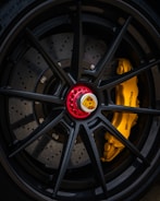 Close-up of a high-performance car wheel featuring a black alloy rim with a red center lock and the Porsche emblem. Behind the spokes, the yellow brake caliper with the Porsche logo is visible, highlighting the precision engineering and luxury design.