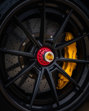 Close-up of a high-performance car wheel featuring a black alloy rim with a red center lock and the Porsche emblem. Behind the spokes, the yellow brake caliper with the Porsche logo is visible, highlighting the precision engineering and luxury design.