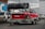 A red sports car is mounted on a metal trailer. Several used tires are stacked on a rack above the car. The scene is set in an urban parking area with other vehicles and buildings visible in the background. The car has numerous decals and is positioned for transportation.