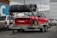 A red sports car is mounted on a metal trailer. Several used tires are stacked on a rack above the car. The scene is set in an urban parking area with other vehicles and buildings visible in the background. The car has numerous decals and is positioned for transportation.
