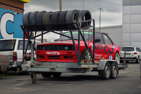 A red sports car is mounted on a metal trailer. Several used tires are stacked on a rack above the car. The scene is set in an urban parking area with other vehicles and buildings visible in the background. The car has numerous decals and is positioned for transportation.