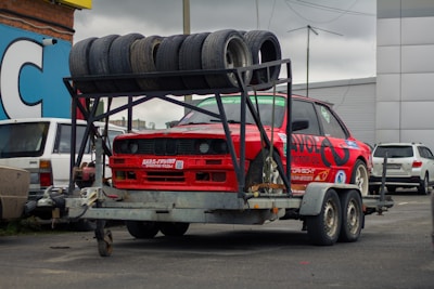 A red sports car is mounted on a metal trailer. Several used tires are stacked on a rack above the car. The scene is set in an urban parking area with other vehicles and buildings visible in the background. The car has numerous decals and is positioned for transportation.