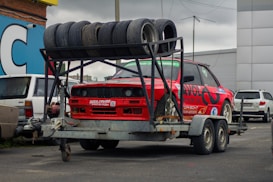 A red sports car is mounted on a metal trailer. Several used tires are stacked on a rack above the car. The scene is set in an urban parking area with other vehicles and buildings visible in the background. The car has numerous decals and is positioned for transportation.
