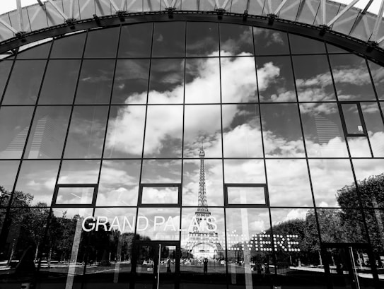 The image captures a reflective glass facade with a mirror-like surface that displays a clear reflection of the Eiffel Tower. The skyline is filled with scattered clouds against a bright sky. The words 'GRAND PALAIS' are visible on the glass, which seems to be part of a modern architectural structure.