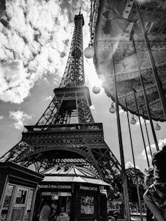 A black and white photograph featuring the Eiffel Tower, with sunlight streaming dramatically through its structure. In the foreground, part of a carousel with spherical lights is visible, providing a contrast to the iconic landmark. Below the tower, there are small kiosks, one with signage in French, adding to the Parisian atmosphere. Fluffy clouds scatter across the sky, enhancing the dynamic composition.