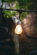 A single light bulb is suspended outdoors, glowing warmly against a blurred background of greenery and brick. The filament inside the bulb is visible, casting a soft, ambient light.