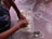 A person is mixing or pressing flour in a rectangular transparent container on a table. The table is covered with a patterned tablecloth featuring heart shapes. A metallic spoon and an empty glass container are nearby. The person is wearing a dark-blue shirt with floral patterns.