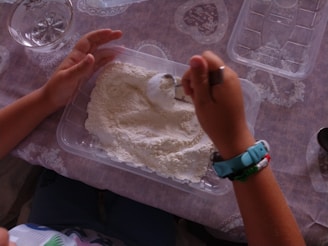 A pair of hands is scooping white powder from a plastic container using a spoon. The setting includes a lace-patterned tablecloth and various plastic containers and dishes are visible in the surroundings. The hands are adorned with colorful bracelets, and the person seems to be engaged in some form of cooking or baking activity.