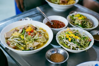 A selection of dishes featuring a variety of fresh ingredients. Bowls of soup are garnished with vibrant vegetables, herbs, and slices of meat. Plates of salad containing sprouts and greens are also present, accompanied by small bowls of dipping sauce. The setting suggests a casual dining environment.