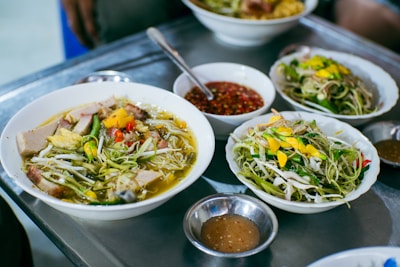 A selection of dishes featuring a variety of fresh ingredients. Bowls of soup are garnished with vibrant vegetables, herbs, and slices of meat. Plates of salad containing sprouts and greens are also present, accompanied by small bowls of dipping sauce. The setting suggests a casual dining environment.