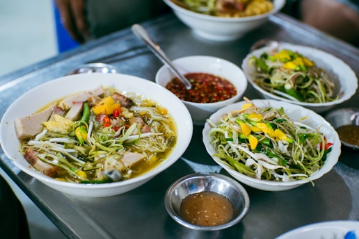 A selection of dishes featuring a variety of fresh ingredients. Bowls of soup are garnished with vibrant vegetables, herbs, and slices of meat. Plates of salad containing sprouts and greens are also present, accompanied by small bowls of dipping sauce. The setting suggests a casual dining environment.
