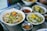 A selection of dishes featuring a variety of fresh ingredients. Bowls of soup are garnished with vibrant vegetables, herbs, and slices of meat. Plates of salad containing sprouts and greens are also present, accompanied by small bowls of dipping sauce. The setting suggests a casual dining environment.