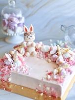 A pastel-colored cake is adorned with decorative flowers and meringues, featuring a cute bunny figurine sitting atop. Additional floral and butterfly decorations surround the edge, enhancing its whimsical design. In the background, a glass jar filled with more pastel-colored confections can be seen, adding to the dreamy, sweet-filled aesthetic.