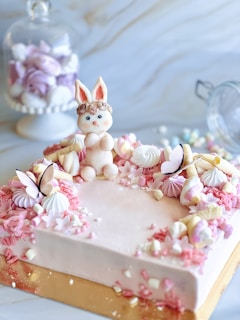 A pastel-colored cake is adorned with decorative flowers and meringues, featuring a cute bunny figurine sitting atop. Additional floral and butterfly decorations surround the edge, enhancing its whimsical design. In the background, a glass jar filled with more pastel-colored confections can be seen, adding to the dreamy, sweet-filled aesthetic.