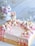 A pastel-colored cake is adorned with decorative flowers and meringues, featuring a cute bunny figurine sitting atop. Additional floral and butterfly decorations surround the edge, enhancing its whimsical design. In the background, a glass jar filled with more pastel-colored confections can be seen, adding to the dreamy, sweet-filled aesthetic.