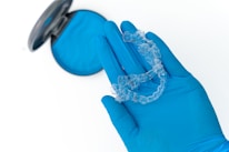 A gloved hand is holding a transparent dental aligner with a dental case in the background. The glove and case are both blue, and the aligner appears to be made of clear plastic.