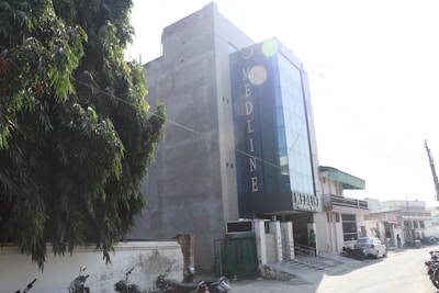 A modern building with a glass facade labeled 'Medline' is situated next to a tree with dense foliage. Several parked motorcycles and a car are visible along the street in front of the building. The urban setting includes additional structures with balconies and awnings.