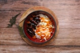 A bowl of rice topped with grilled meat, shredded cabbage, and sauces is placed on a round wooden board. A wooden spoon rests next to the bowl. The background features a wooden surface with fern-like greenery.
