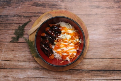 A bowl of rice topped with grilled meat, shredded cabbage, and sauces is placed on a round wooden board. A wooden spoon rests next to the bowl. The background features a wooden surface with fern-like greenery.