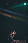A DJ stands at a console with headphones around his neck, illuminated by colorful stage lights. He is focused on his work in a dark setting, with green and yellow beams creating a dynamic atmosphere.