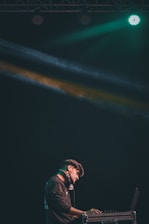 A DJ stands at a console with headphones around his neck, illuminated by colorful stage lights. He is focused on his work in a dark setting, with green and yellow beams creating a dynamic atmosphere.