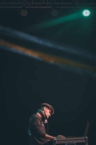 A DJ stands at a console with headphones around his neck, illuminated by colorful stage lights. He is focused on his work in a dark setting, with green and yellow beams creating a dynamic atmosphere.
