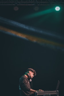 A DJ stands at a console with headphones around his neck, illuminated by colorful stage lights. He is focused on his work in a dark setting, with green and yellow beams creating a dynamic atmosphere.