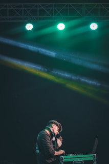 A DJ is performing on stage under vibrant green lighting. He is wearing headphones and appears focused as he works at the DJ console.