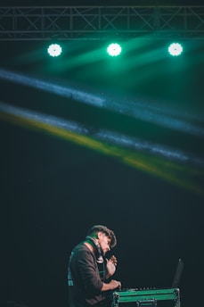 A DJ is performing on stage under vibrant green lighting. He is wearing headphones and appears focused as he works at the DJ console.