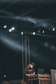 A person is performing on stage with a laptop and DJ equipment, under a framework of lights and cables. The scene is dark with bright spotlights creating beams of light and some haze filling the area. The person appears focused on their equipment.