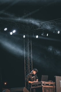 A person is performing on stage with a laptop and DJ equipment, under a framework of lights and cables. The scene is dark with bright spotlights creating beams of light and some haze filling the area. The person appears focused on their equipment.